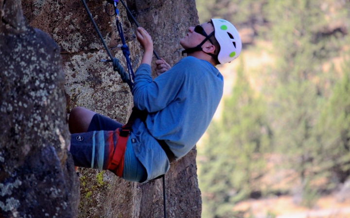 Wearing safety gear and secured by ropes, a person climbs a rock wall. There are trees in the distance in the background. 